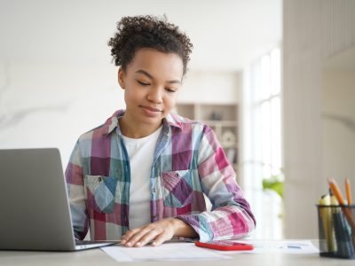 Mixed race teen student girl learning sitting at desk with laptop at home, preparing for school test